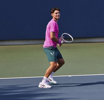 Mannelijke tennisser in roze shirt en groene shorts bereidt zich voor om de bal te slaan op een buitenbaan met blauwe en groene oppervlakken.