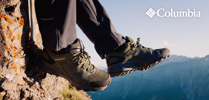 Schwarze und olivgrüne Wanderschuhe mit einem Obermaterial aus Mesh, einer dicken Gummisohle und einem robusten Profil, die an einer unebenen Oberfläche am Rand eines Berges lehnen.