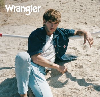 Homme aux cheveux en désordre portant une veste en jean foncé et un jean clair, allongé sur un sol sablonneux près d'un poteau horizontal blanc et rouge.