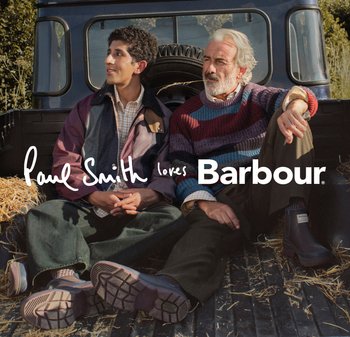 Two men sit on a vehicle's tailgate in a field. One wears a multi-colored striped sweater; the other, a purple jacket over a checkered shirt.