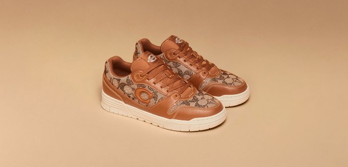 Coppia di sneakers marroni e beige con motivo pattern e suole bianche, posate su una superficie beige, che mostrano il design con lacci e dettagli con logo brandizzato.