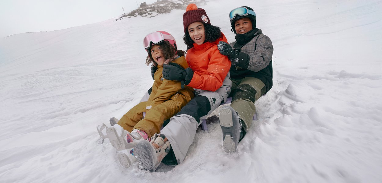 Trys vaikai sėdi rogutėse sniege. Jie dėvi spalvotus slidinėjimo švarkus: oranžinį, rudą ir pilką. Ant jų yra šalmai, jie šypsosi, apsupti sniego.