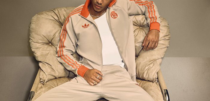 Άνδρας με μπεζ φόρμα Adidas με πορτοκαλί ρίγες κάθεται άνετα σε μια μπεζ μαξιλαρομένη καρέκλα μπροστά σε ουδέτερο φόντο.