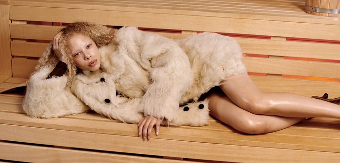 Cappotto in pelliccia sintetica color crema, con bottoni neri, indossato su abbigliamento scuro. La modella si rilassa su una panchina di legno con texture liscia e tonalità calde.