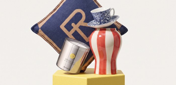 Coussin décoratif bleu et or, bougie parfumée argentée, vase rayé rouge et blanc, et tasse et soucoupe à thé à motif bleu et blanc sur un piédestal jaune.