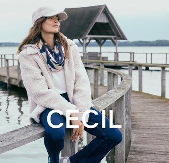 Flauschige rosa Jacke, weißer Pullover, blaue Jeans, gemusterter Schal und graue Mütze. Model sitzt auf einem Holzsteg an einem Gewässer.