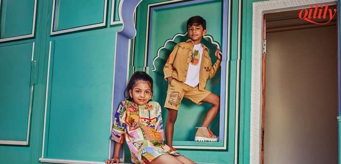 Twee kinderen poseren tegen een turquoise en witte architecturale achtergrond; meisje draagt een kleurrijke jurk, jongen draagt een beige jas en korte broek met patchwork.