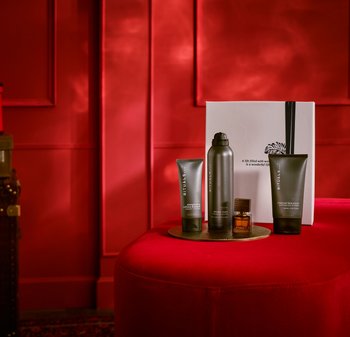 Fire skincare produkter arrangert på en rund gullfat: grå rør, en metallbeholder, og en liten glassflaske, satt mot en rød bakgrunn.