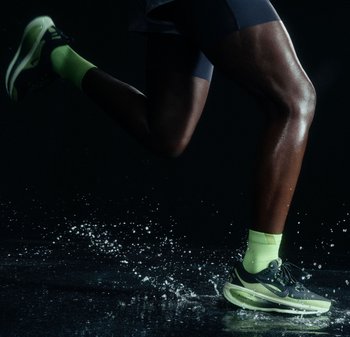 Gambe di un corridore in movimento con scarpe e calze verdi brillanti che schizzano acqua su una superficie scura con sfondo nero.
