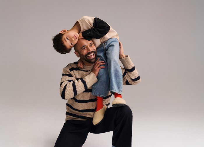 Άνδρας γονατιστός με παιδί που ακουμπάει ανάποδα στους ώμους του, και οι δύο χαμογελούν με φόντο απλό γκρι.