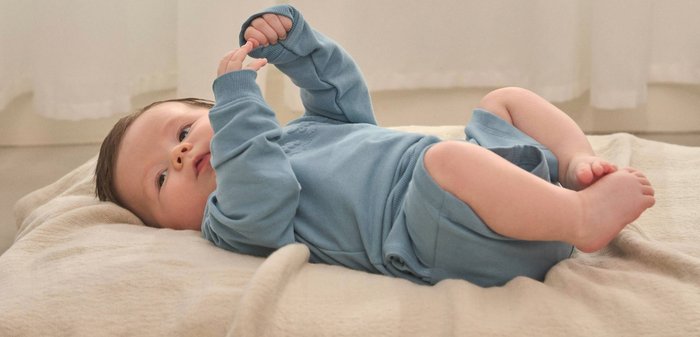 Lächendes Baby liegt auf beigem Stoff, trägt ein cremefarbenes Langarmshirt mit Segelboot- und Leuchtturm-Motiv und blaue Hose.