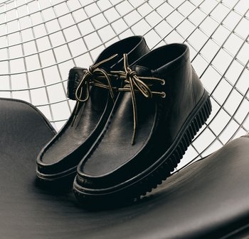 Chaussures en cuir noir avec un design à cheville basse, ornées de lacets dorés et d'une semelle en caoutchouc striée, posées sur une surface noire avec un fond en fil.