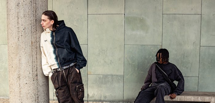 Dva jednotlivci stojí proti texturované kamenné zdi. Jeden má na sobě černobílé mikino s rozdělenou kapucí a druhý je oblečen v celém černém oděvu, sedí.