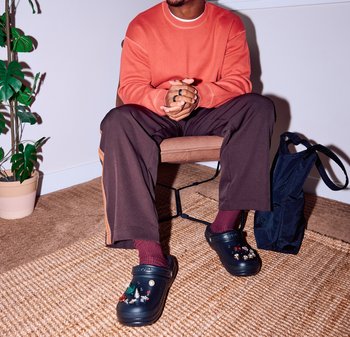 Črne klompe z okrasnimi čarobnimi elementi, oranžna pulover, rjave hlače širokega kroja, kostanjeve nogavice, sedeč na stolu, s temno torbo v bližini.