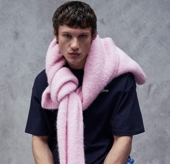 Giovane con capelli corti e ricci che indossa una grande sciarpa rosa pelosa drappeggiata sulle spalle e una maglietta blu scuro su sfondo grigio.