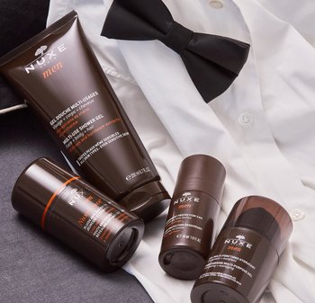 Il set di cura NUXE Men include un tubetto di gel doccia marrone, tre barattoli per la cura della pelle marroni e un papillon nero su una camicia bianca.