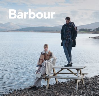 Two models by a lake wearing Barbour clothing. One wears a beige and brown checkered poncho; the other has a dark coat and scarf.