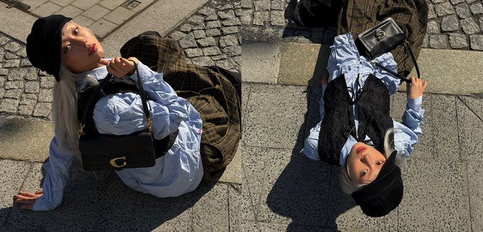 Persona con capelli chiari che indossa un basco nero, un gilet nero, una camicetta blu e pantaloni a quadri, tenendo una borsa nera mentre è seduta e sdraiata sul marciapiede.