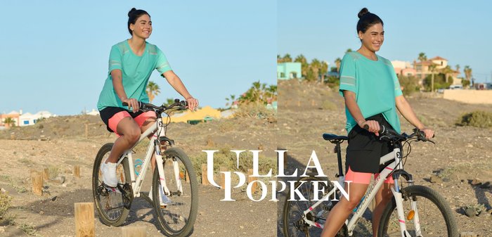 Young woman in teal shirt and coral shorts riding a white mountain bike on a dirt path with scattered wooden posts.