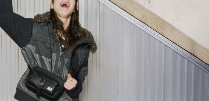 Person trägt Sonnenbrille, dunkle Jacke mit Pelzkragen und Umhängetasche, lächelt und hebt einen Arm drinnen vor einer Paneelwand.