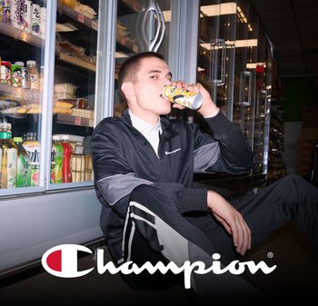 Black athletic tracksuit with gray accents, zippered jacket, and stripes on pants. Person sipping from a colorful can. Refrigerated display in background.