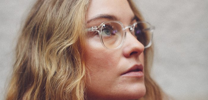 Vrouw met lang, golvend blond haar en een bril met doorzichtige montuur, die omhoog kijkt tegen een neutrale achtergrond.