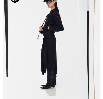 Donna con giacca nera, gonna asimmetrica sopra i pantaloni, stivali a punta e occhiali da sole neri futuristici, in piedi di lato contro uno sfondo bianco.