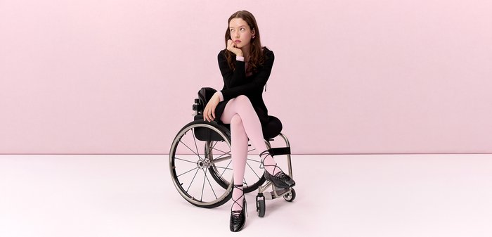 Young woman in black dress and pink tights sitting thoughtfully in a wheelchair against a soft pink background.