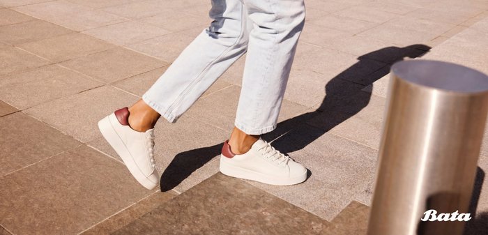 Person, die auf einem gepflasterten Gehweg läuft, trägt hellblaue Jeans und weiße Turnschuhe mit weinroten Akzenten.