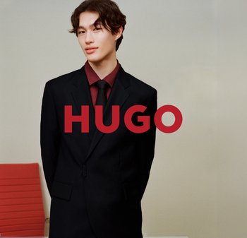 Costume noir avec revers en encoche, porté sur une chemise bordeaux et une cravate assortie. L'arrière-plan présente des couleurs claires et une chaise rouge.