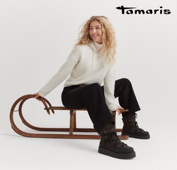 Weißer Kapuzenpulli aus weichem Material; schwarze, weit geschnittene Hose; gemusterte Stiefel mit dicker Plattform; sitzt auf einem Holzschlitten.