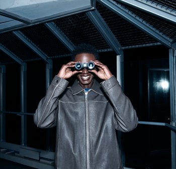 Junger Mann in grauer Jacke lächelt, während er nachts in einem dunklen, metallgerahmten Korridor schwarze Ferngläser an die Augen hält.