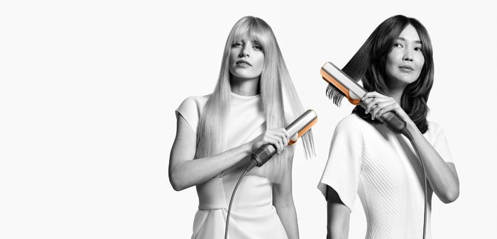 Haarstylingset met een metallic zilveren behuizing en oranje accenten, vastgehouden door twee individuen met lang, steil haar in contrasterende stijlen.