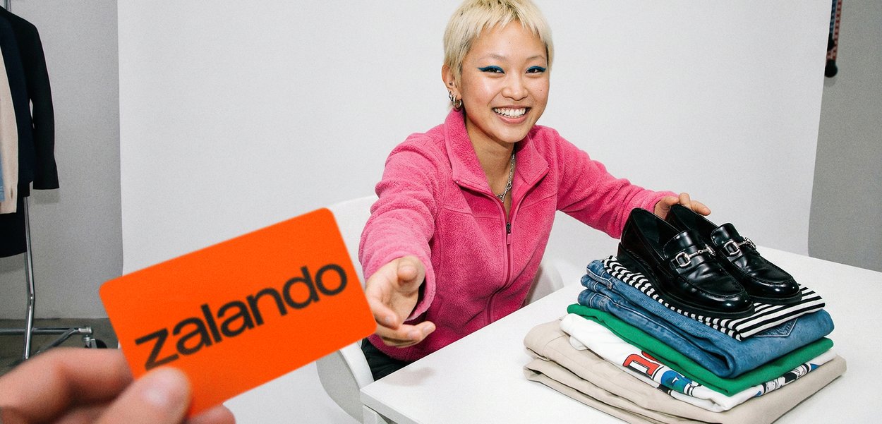 Lachend persoon in een roze jas die reikt naar een feloranje Zalando-kaart bij een tafel met opgevouwen kleding en zwarte nette schoenen.