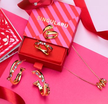 Set di gioielli tono oro comprendente due paia di orecchini a cerchio con pietre colorate, un anello con design floreale e una collana coordinata in una scatola rossa.