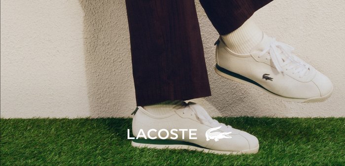 Osoba sediaca na bielom okraji pri oplotenom plote, oblečená v tmavých nohaviciach a bielych teniskách Lacoste na zelenej tráve. Logo Lacoste je viditeľné.
