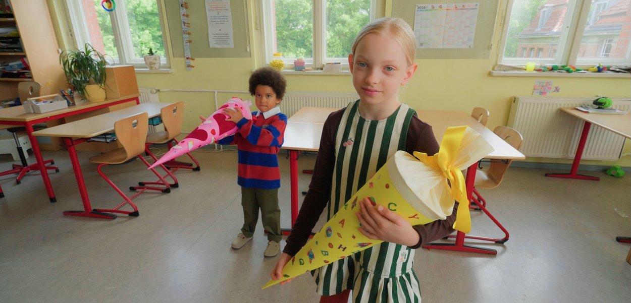 Zwei Kinder stehen in einem Klassenzimmer und halten große bunte kegelförmige Schulgaben, die mit Bändern verpackt sind.