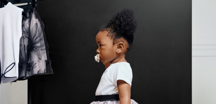 Bambina piccola con ciuccio, indossa una maglietta bianca e una gonna di tulle a fantasia, in piedi di profilo contro uno sfondo nero vicino a vestiti appesi.