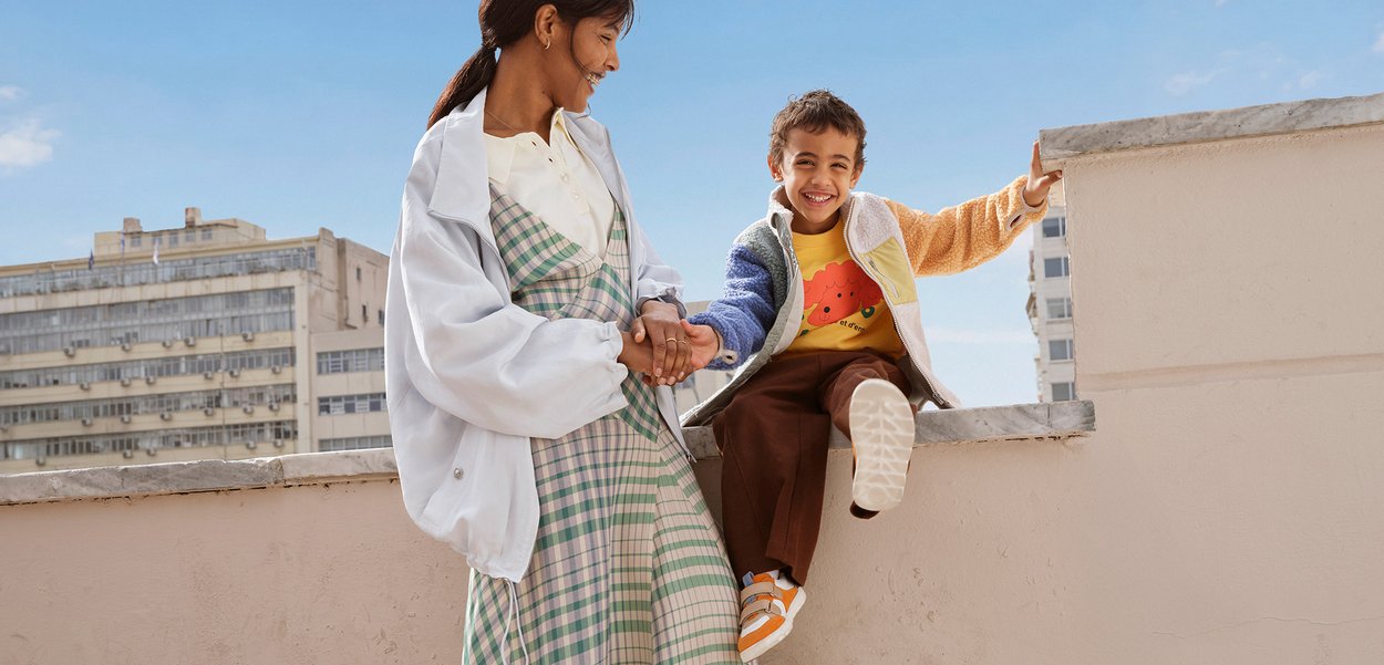 Mujer tomándose de la mano con niño sonriente sentado en el borde de un tejado con cielo azul y edificios urbanos de fondo.