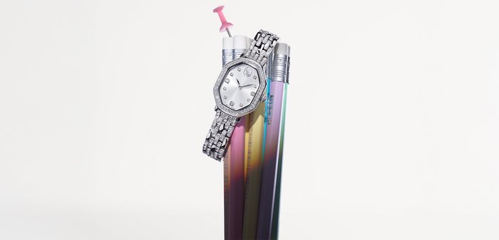 Silberner achteckiger Uhr mit Diamantakzenten an einem Metallarmband, angeordnet unter bunten Buntstiften in abgestuften Farben vor einem schlichten Hintergrund.