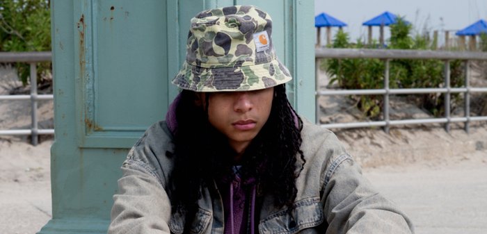 Sombrero tipo bucket de camuflaje en tonos de verde y marrón, material de algodón. Se lleva con una chaqueta de mezclilla gris sobre un sudadera morada.