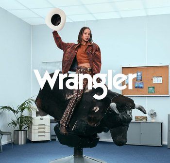 Person med brun jakke og mønstrede bukser rir på mekanisk okse innendørs, holder opp en hvit hatt, med kontorbakgrunn og Wrangler-logo.
