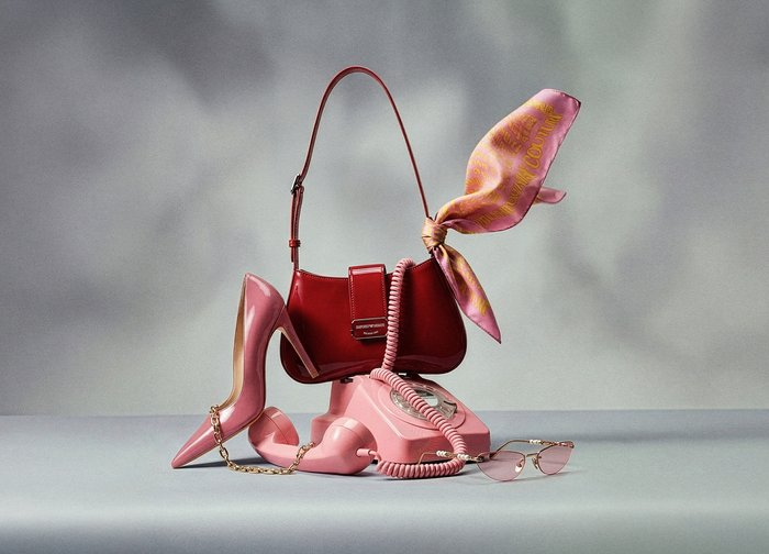 Téléphone rotatif rose avec un sac à main rouge attaché au combiné par une écharpe rose, chaussure à talon haut rose et lunettes de soleil teintées de rose sur une surface grise.