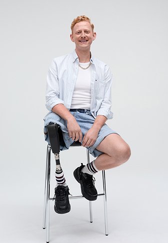 Una persona con una camicia a righe blu chiaro e una canotta bianca è seduta su uno sgabello d'argento, indossando pantaloni shorts in denim e calze a righe, con una gamba prostetica.