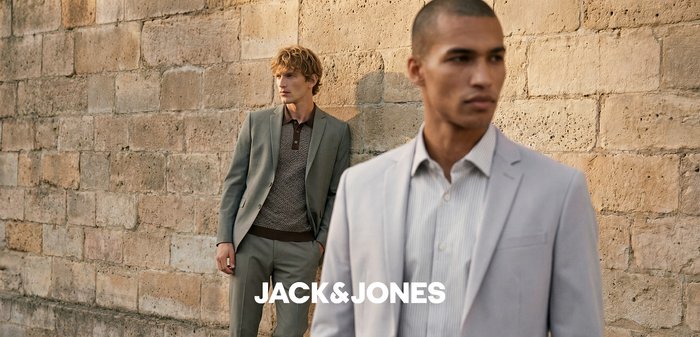 Dois homens estão junto a uma parede de pedra com textura, um veste um fato cinzento com um suéter estampado, o outro um blazer claro e uma camisa às riscas.