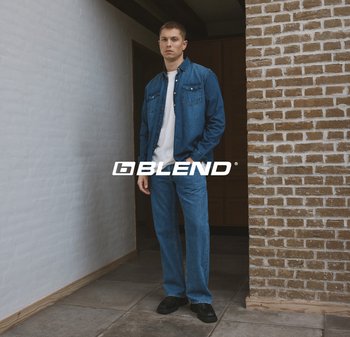 Model draagt een spijkerjasje en bijpassende spijkerbroek in een medium blauwe tint, gecombineerd met een wit T-shirt en zwarte schoenen. Achtergrond van bakstenen muur.