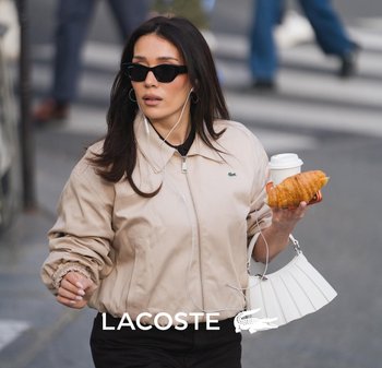 Mujer con chaqueta beige de Lacoste y gafas de sol negras, sosteniendo un croissant y una taza de café mientras camina por una calle de la ciudad.