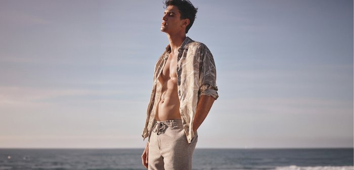 Young man with open patterned shirt and shorts standing with eyes closed by the ocean under a clear sky.