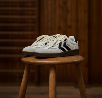 Witte suède sneakers met donkerblauwe zigzagaccenten, ronde neus, rubberen zool en beige veters, tentoongesteld op een houten kruk.