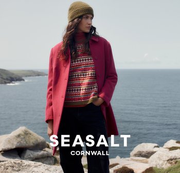 Červený kabát s texturovaným povrchem, vzorovaný pletený svetr, zelená beretka a tmavé kalhoty na pozadí pobřeží.